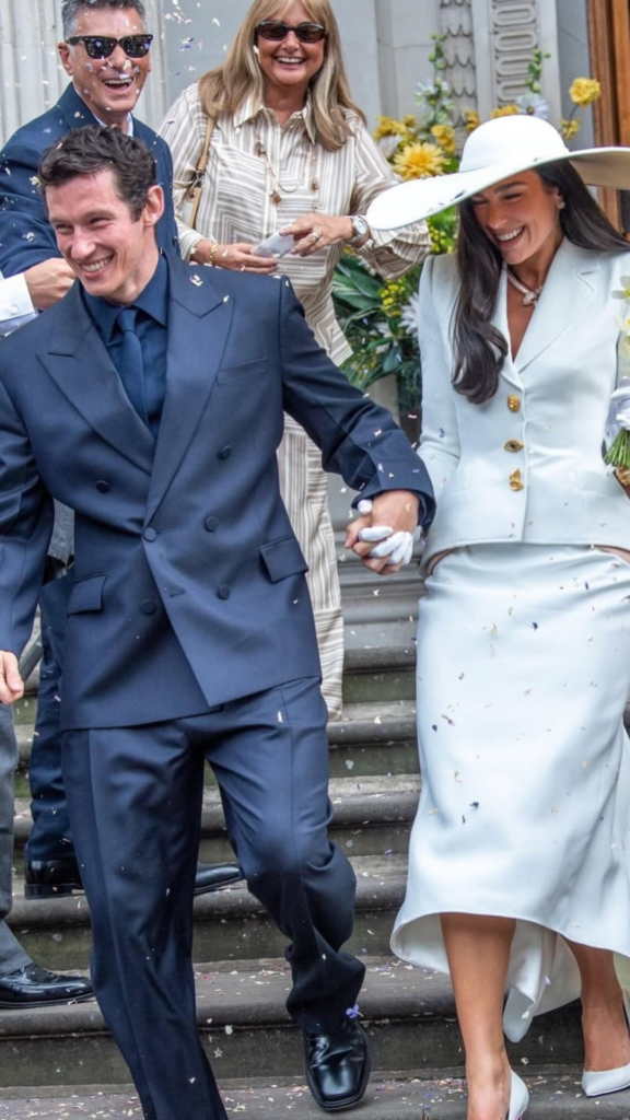 Dua Lipa smiling in white Schiaparelli skirt suit and Stephen Jones hat outside London registry office